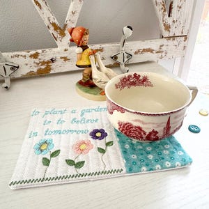 Pu&ograve; includere: Un sottobicchiere ricamato bianco con un motivo floreale e la scritta "to plant a garden is to believe in tomorrow". Una grande tazza di ceramica con un motivo rosso e bianco &egrave; appoggiata sul sottobicchiere. Statuine decorative sullo sfondo.
