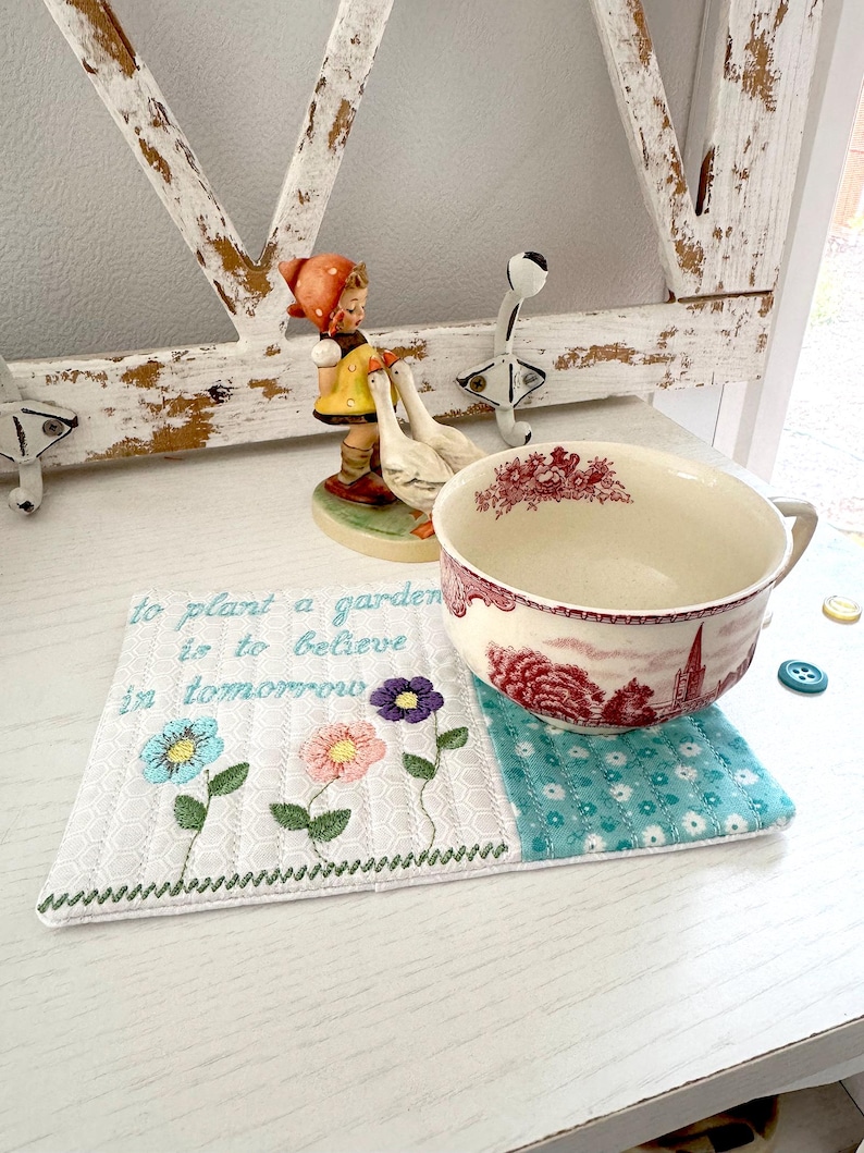 Pu&ograve; includere: Un sottobicchiere ricamato bianco con un motivo floreale e la scritta "to plant a garden is to believe in tomorrow". Una grande tazza di ceramica con un motivo rosso e bianco &egrave; appoggiata sul sottobicchiere. Statuine decorative sullo sfondo.