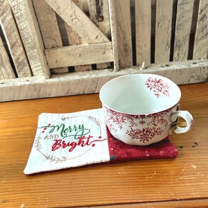 May include: A white and red embroidered coaster with the text "Merry and Bright" in green and red lettering. The coaster is sitting on a wooden surface with a white teacup with a red floral pattern on top of it.