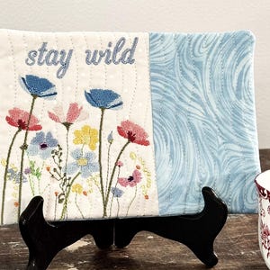 Puede incluir: Un panel de tela rectangular con flores bordadas y las palabras "stay wild". El lado izquierdo presenta flores coloridas, mientras que el lado derecho tiene un patrón de ondas azules. El panel se exhibe en un soporte negro.