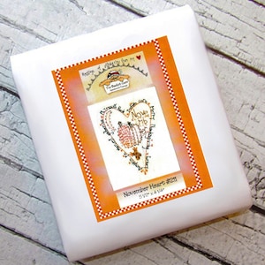 Op de afbeelding: Oranje en wit hartvormig borduurpatroon met de tekst "Plymouth Rock Indian corn Harvest gathering Pumpkin Harvest Thanksgiving Turkey Nove Mb" en "November Heart #211 5 1/2" x 4 1/4"".