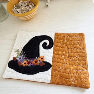 May include: A rectangular quilted fabric coaster featuring a white side with a black witch hat adorned with colorful embroidered flowers and a mustard yellow side with a stitched star pattern. The coaster has a soft, textured surface.