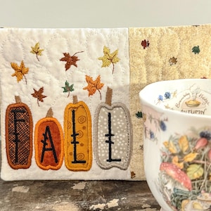 May include: A quilted fabric mat featuring four embroidered pumpkins in brown, orange, yellow, and grey, each displaying a letter to spell the word FALL. The mat is decorated with scattered embroidered autumn leaves and sits beside a ceramic mug.