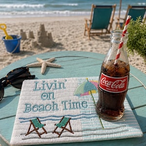 May include: A rectangular fabric mat featuring embroidered text that reads Livin' on Beach Time, accompanied by stitched designs of two green beach chairs and a colorful umbrella, placed on a round table with a glass bottle of Coca-Cola and a straw.