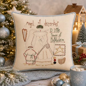 May include: A square cream-coloured throw pillow featuring intricate embroidery of an angel, a shepherd, a star, and a heart. The text reads Angel of december and O Little town of Bethlehem. The pillow has a light blue piped edge and three small buttons.