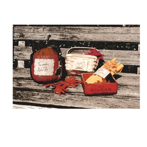 Op de afbeelding: Een rustieke houten bank met drie herfstthemadecoraties: een kleine rode en bruine pompoen met de tekst "Tiny pumpkins on a porch do", een witte rieten mand met de tekst "Pumpkins" en een rode houten doos met de tekst "Chelsea Clock Co. Boston, Mass." met gele kaarsen.