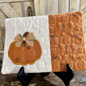 May include: A rectangular quilted fabric coaster featuring a split design. The left side is white with an embroidered orange pumpkin topped with a tan bow. The right side is solid orange with a repeating quilted pumpkin and star pattern.