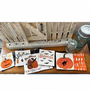 Peut inclure: Cinq dessous-de-plat matelassés sur le thème d'Halloween avec des motifs de citrouilles et de chauves-souris. Les dessous-de-plat portent les inscriptions "jack", "Spookie", "October", "Halloween" et "TRICKS SBOO TRE CARY".