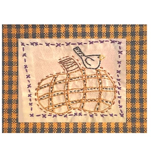 May include: A hand-stitched embroidery design featuring a brown pumpkin with a small blue bird perched on top. The design is framed with a purple stitched border and set against a yellow and black gingham background.