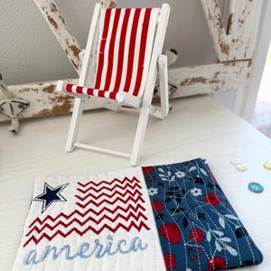 Pu&ograve; includere: Una sedia a sdraio in legno bianco in miniatura con tessuto a righe rosse e bianche. Un sottobicchiere trapuntato con un disegno della bandiera americana ricamato e la parola "America" in blu. Il sottobicchiere ha un motivo floreale sul lato destro.
