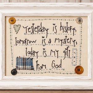 May include: A white distressed wooden frame containing a beige fabric panel with black embroidered text that reads: Yesterday is history, Tomorrow is a mystery, today is my gift from God. The panel features four buttons and a blue plaid fabric patch.