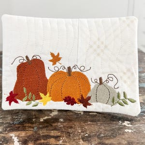 May include: A rectangular quilted fabric piece in off-white featuring machine embroidery of three pumpkins in burnt orange, bright orange, and muted sage green. The design includes small embroidered autumn leaves in red, yellow, and brown.