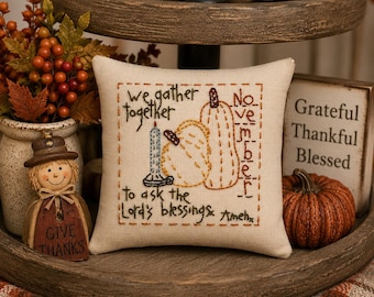 May include: A square beige fabric pillow featuring brown embroidered text that reads We gather together to ask the Lord's blessings Amen and November. The design includes an embroidered blue candle and two yellow pumpkins outlined in brown thread.
