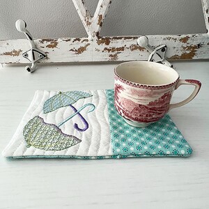 May include: A white quilted coaster with an embroidered design of two umbrellas in shades of green, purple, and blue. The coaster is paired with a red and white teacup with a floral pattern.