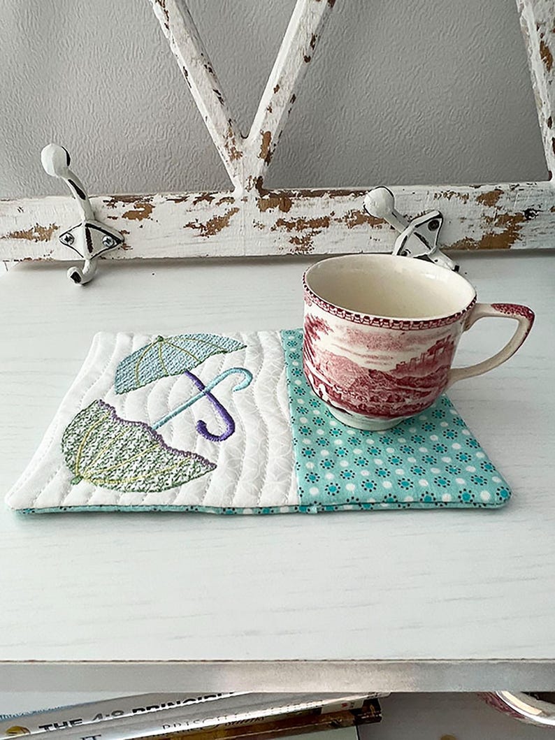 May include: A white quilted coaster with an embroidered design of two umbrellas in shades of green, purple, and blue. The coaster is paired with a red and white teacup with a floral pattern.