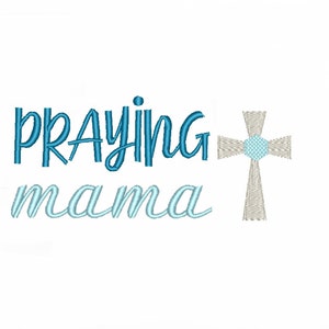 May include: An embroidered design featuring the words "PRAYING mama" in blue and light blue script. A cross with a light blue center and tan cross arms is to the right of the text. The design is on a white background.
