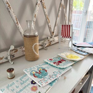 Pu&ograve; includere: Una composizione decorativa con una bottiglia di vetro con una custodia in juta e un design a cuore, una sedia in miniatura a strisce rosse e bianche e diversi sottobicchieri ricamati. I sottobicchieri presentano motivi floreali e a tema spiaggia, con il testo "Life is better in Flip Flops".
