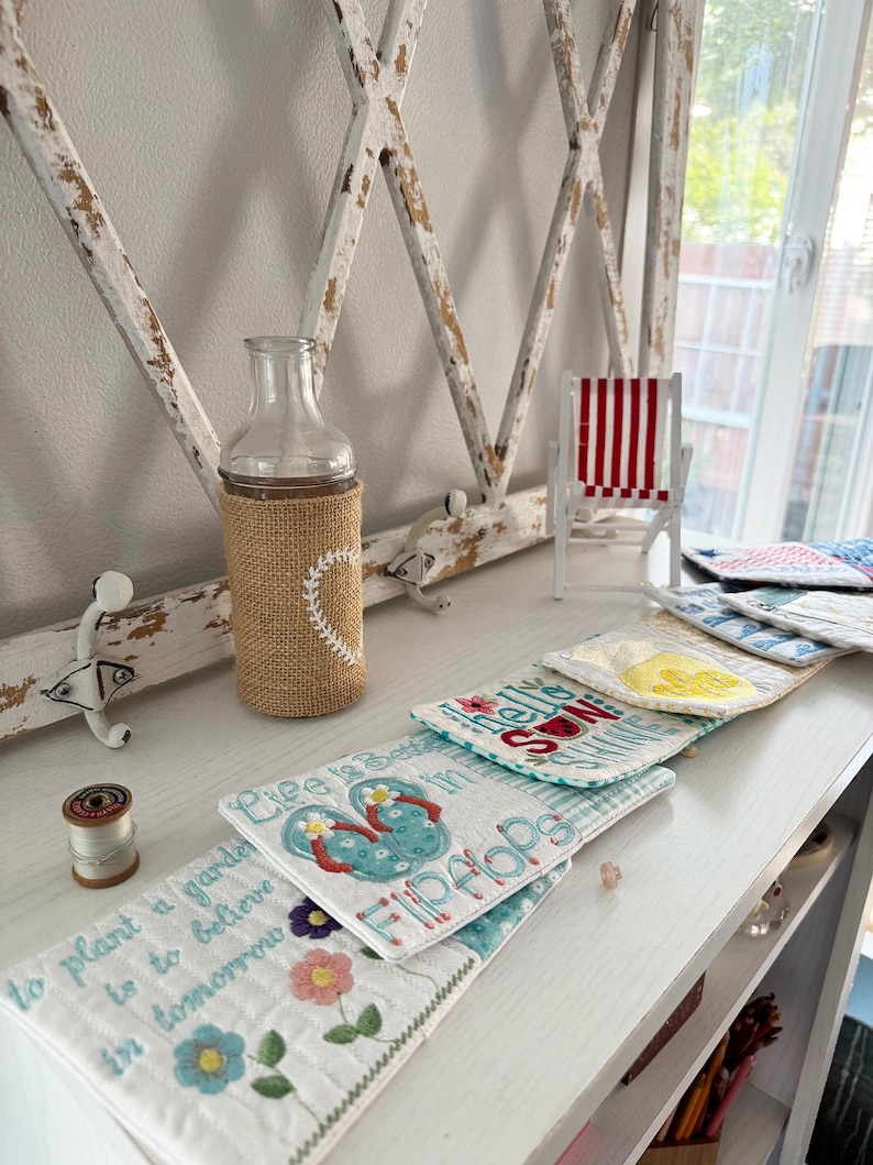 Pu&ograve; includere: Una composizione decorativa con una bottiglia di vetro con una custodia in juta e un design a cuore, una sedia in miniatura a strisce rosse e bianche e diversi sottobicchieri ricamati. I sottobicchieri presentano motivi floreali e a tema spiaggia, con il testo "Life is better in Flip Flops".