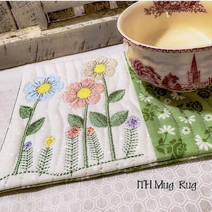 Può includere: Sottobicchiere ricamato con fiori colorati su sfondo bianco. Il tappeto ha un bordo verde e una sezione a motivi floreali verdi. Una ciotola con motivi floreali è appoggiata sul tappeto. Il testo "MH Mug Rug" è visibile.
