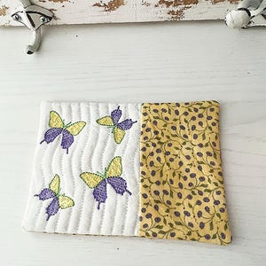 May include: A white quilted coaster with a yellow and green floral pattern on one side and four embroidered purple butterflies on the other side.