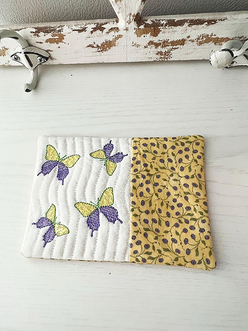 May include: A white quilted coaster with a yellow and green floral pattern on one side and four embroidered purple butterflies on the other side.