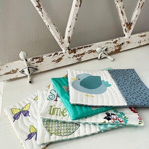 May include: A stack of five quilted fabric coasters with various designs. The top coaster features a blue bird, the second coaster says "Springtime", the third coaster has a butterfly design, the fourth coaster has an umbrella design, and the bottom coaster has a floral design.