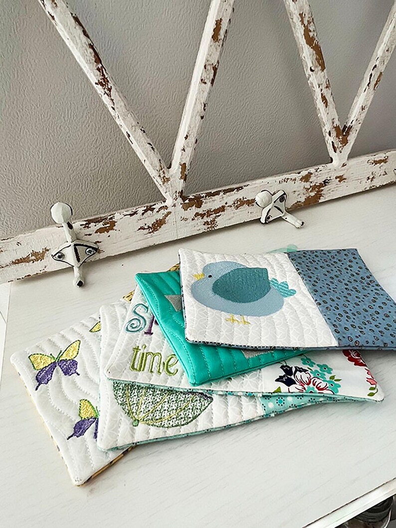 May include: A stack of five quilted fabric coasters with various designs. The top coaster features a blue bird, the second coaster says "Springtime", the third coaster has a butterfly design, the fourth coaster has an umbrella design, and the bottom coaster has a floral design.