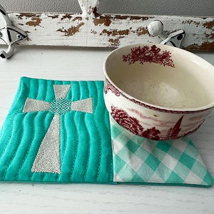 May include: A teal blue quilted coaster with an embroidered white cross design. The coaster is sitting on a white surface with a white and red patterned bowl on top of it.