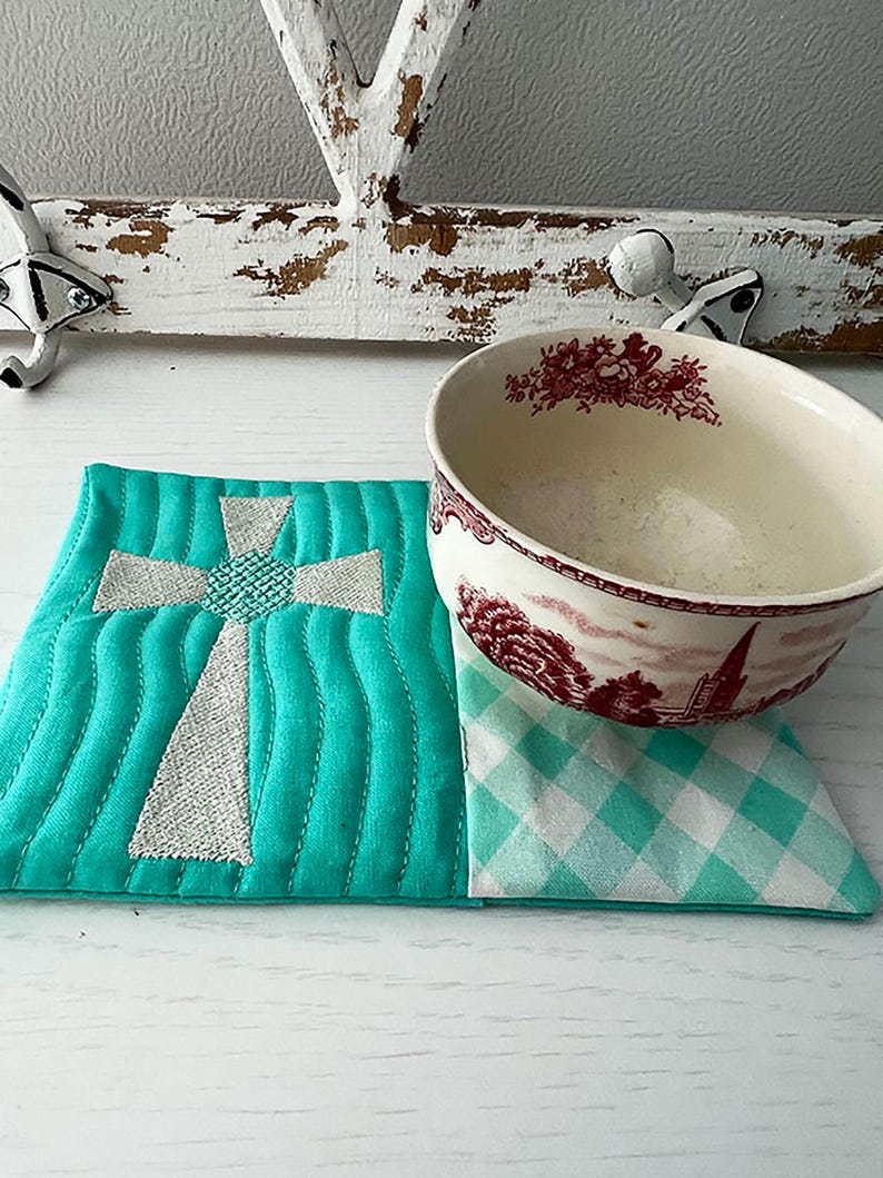 May include: A teal blue quilted coaster with an embroidered white cross design. The coaster is sitting on a white surface with a white and red patterned bowl on top of it.