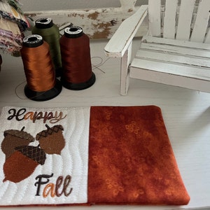 Pode incluir: Um descanso de copo acolchoado branco com o texto "Happy Fall" e um desenho bordado de três bolotas. O descanso de copo é combinado com um tecido castanho com um padrão branco.