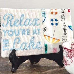 May include: A white quilted lake-themed sign with blue embroidered text that reads "Relax You're at the Lake". The sign is propped up on a black stand. The right side of the sign has a pattern of life preservers, umbrellas, and the text "all I need is a little Vit".