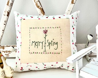 May include: A white pillow with a pink floral pattern and a cream-colored embroidered patch that says "Merry Spring" with a green stem and red flower.