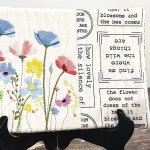 Könnte beinhalten: Eine rechteckige, gesteppte Stofftasche mit gestickten Wildblumen in Blau, Rosa und Gelb auf weißem Grund. Die rechte Seite zeigt gedruckten schwarzen Text, darunter Phrasen wie find me where the wild things are.