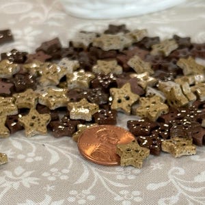 May include: A collection of star-shaped buttons in shades of brown, tan, and beige. The buttons have four holes for sewing and are scattered on a patterned surface. A U.S. penny is included for size reference.