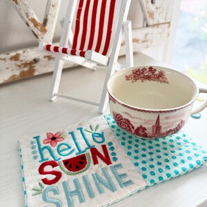 Pu&ograve; includere: Una sedia a sdraio bianca in miniatura con tessuto a righe rosse e bianche. Una tazza da t&egrave; con un disegno floreale rosso &egrave; appoggiata su un sottobicchiere trapuntato bianco e blu a pois con le parole "hello sunshine" ricamate.