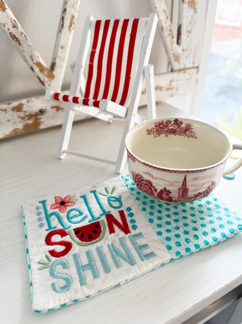 Pu&ograve; includere: Una sedia a sdraio bianca in miniatura con tessuto a righe rosse e bianche. Una tazza da t&egrave; con un disegno floreale rosso &egrave; appoggiata su un sottobicchiere trapuntato bianco e blu a pois con le parole "hello sunshine" ricamate.