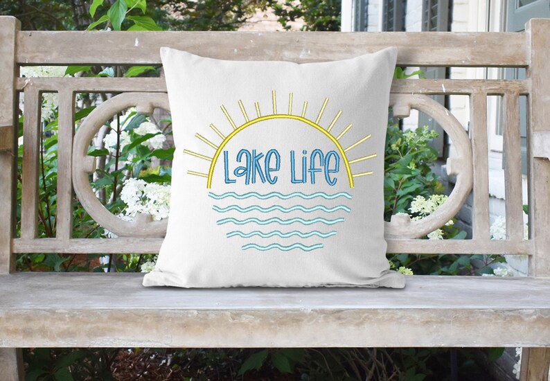 May include: A white square throw pillow featuring a central graphic design of a yellow sun with radiating rays above the text "Lake Life" in blue, positioned over a series of light blue wavy lines representing water, centered on the pillow fabric.