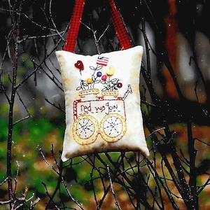 May include: A white fabric pillow with red trim hangs from a branch. The pillow is embroidered with a red wagon carrying flowers and the text "Red wagon".