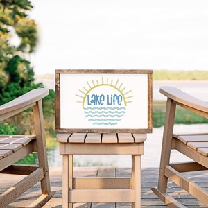 May include: A rectangular wooden sign with a white background featuring the text Lake Life in blue letters. Above the text is a yellow sun graphic with rays, and below the text are blue wavy lines representing water. The sign has a dark wood frame.