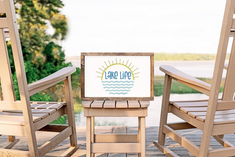 May include: A rectangular wooden sign with a white background featuring the text Lake Life in blue letters. Above the text is a yellow sun graphic with rays, and below the text are blue wavy lines representing water. The sign has a dark wood frame.