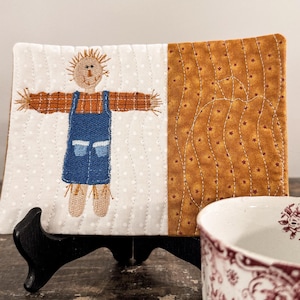 Puede incluir: Un objeto decorativo rectangular acolchado con un diseño de espantapájaros cosido. El espantapájaros tiene una cara marrón, brazos marrones y un peto azul. El lado derecho es de tela marrón dorada con un estampado de estrellas.