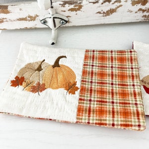 May include: A white and orange embroidered pumpkin design on a white fabric coaster with a plaid fabric backing.