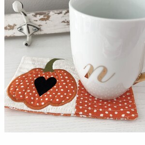 May include: A white ceramic mug with a gold letter "N" on the side, resting on a quilted pumpkin-shaped coaster with white polka dots on an orange background.