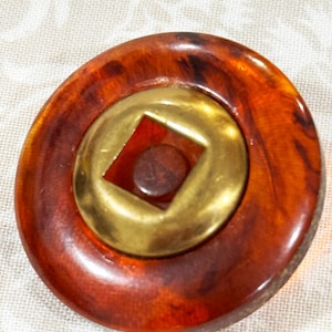 May include: A round, vintage brooch with a tortoiseshell-like pattern in shades of brown and amber. The center features a gold-colored metal circle with a square cut-out and a small, dark brown center. The brooch has a glossy finish.