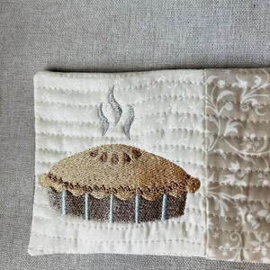 May include: Embroidered fabric square featuring a pie design. The pie has a golden-brown crust and a dark brown filling, with steam rising above it. The background is a quilted cream fabric with a floral pattern.