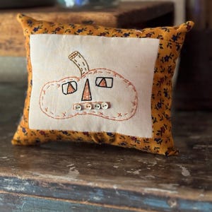 May include: A small, rectangular decorative pillow with a pumpkin design. The pumpkin is embroidered on a white fabric square, with orange stitching and button details. The pillow's border is a yellow floral pattern.