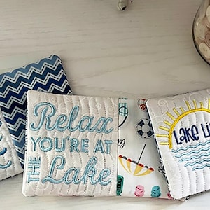 May include: Six white fabric coasters with blue embroidery. The coasters feature lake-themed sayings, including "Lake Hair Don't Care", "Relax You're at the Lake", "Lake Life", and "The Lake is Calling I Must Go".
