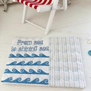 May include: A quilted coaster with a nautical theme. The coaster is divided into two sections: one with blue embroidered waves and the other with vertical stripes. The text "From sea to shining sea" is embroidered in blue. A miniature red and white striped beach chair is in the background.