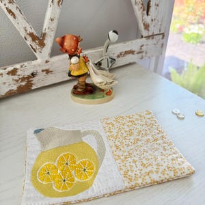 Pu&ograve; includere: Un sottobicchiere in tessuto fatto a mano con un motivo a brocca gialla e fette di limone, su uno sfondo floreale bianco e giallo. Il sottobicchiere &egrave; posto su una superficie bianca, con una statuina decorativa sullo sfondo.