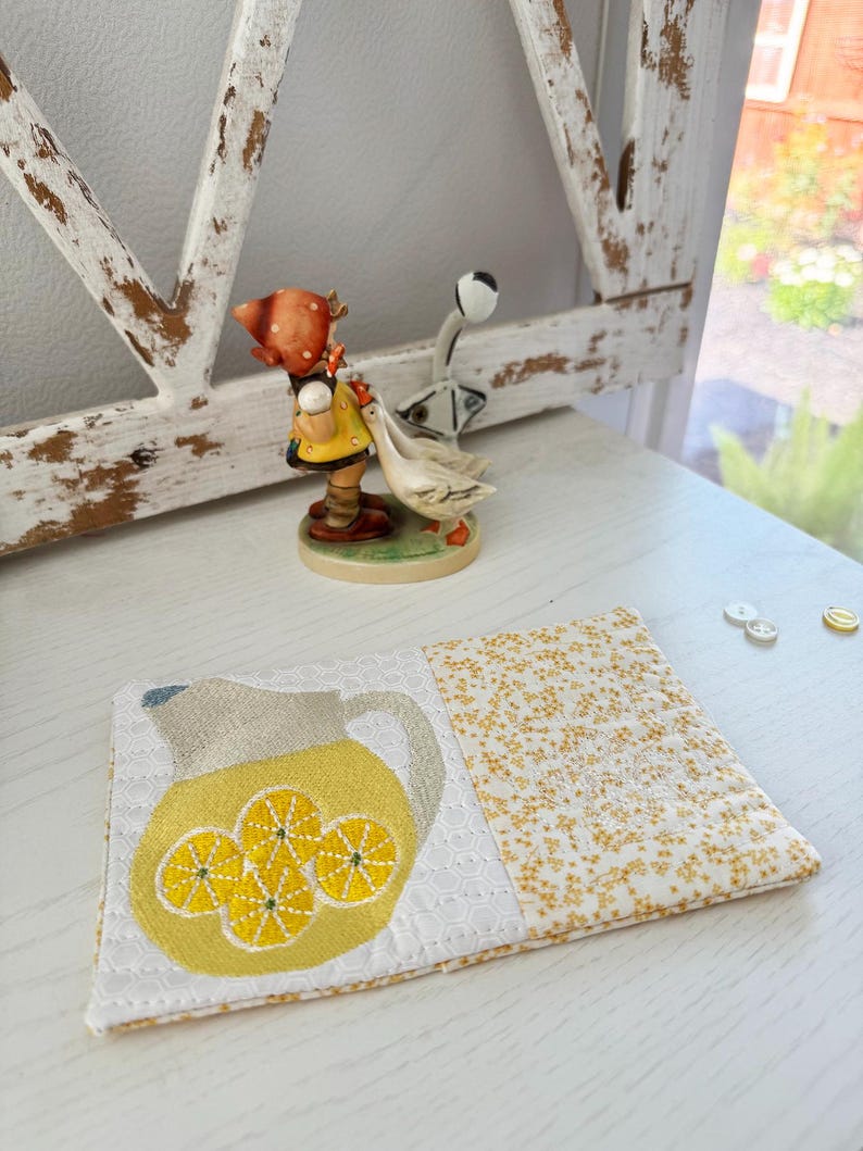 Pu&ograve; includere: Un sottobicchiere in tessuto fatto a mano con un motivo a brocca gialla e fette di limone, su uno sfondo floreale bianco e giallo. Il sottobicchiere &egrave; posto su una superficie bianca, con una statuina decorativa sullo sfondo.